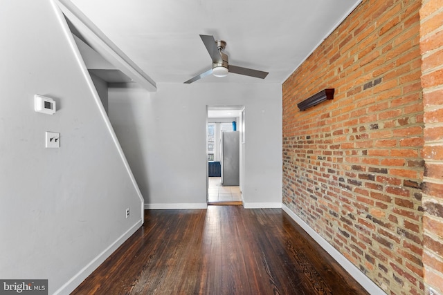 spare room with dark wood-type flooring, baseboards, ceiling fan, and brick wall