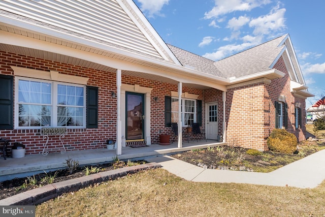 property entrance featuring covered porch, brick siding, and roof with shingles