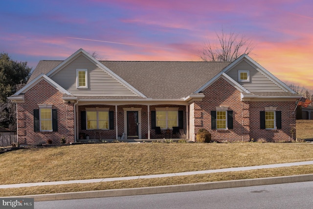 view of front facade with a front yard, covered porch, brick siding, and roof with shingles