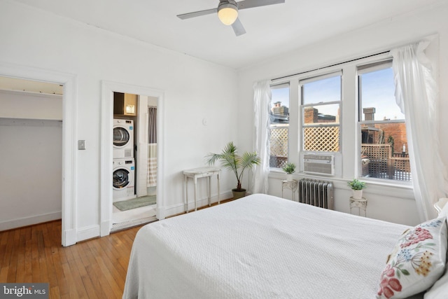 bedroom with wood finished floors, a ceiling fan, baseboards, stacked washer / drying machine, and radiator heating unit