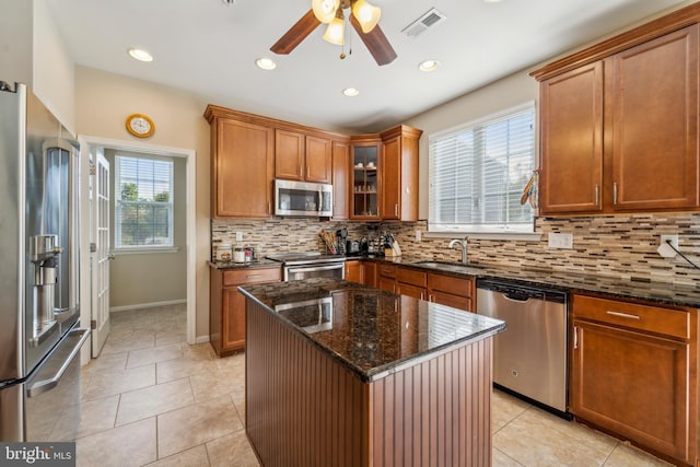 kitchen with appliances with stainless steel finishes, brown cabinetry, glass insert cabinets, a sink, and dark stone countertops