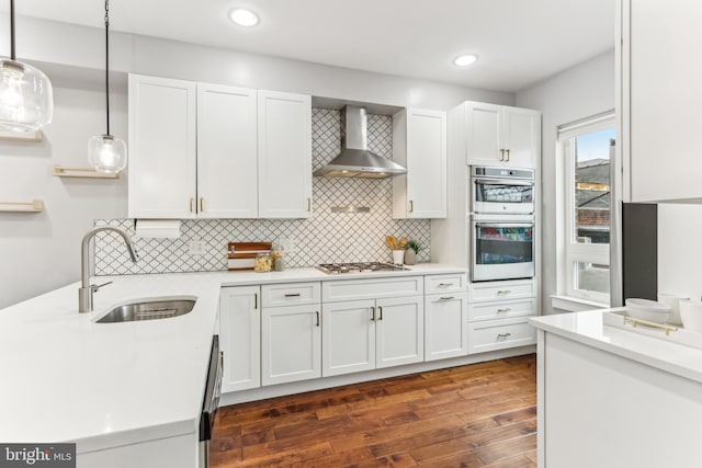 kitchen with stainless steel appliances, a sink, light countertops, wall chimney range hood, and dark wood-style floors