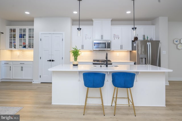 kitchen with a kitchen island with sink, white cabinetry, hanging light fixtures, appliances with stainless steel finishes, and glass insert cabinets