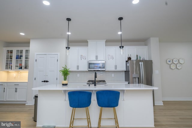 kitchen featuring decorative light fixtures, a center island with sink, stainless steel appliances, light countertops, and glass insert cabinets