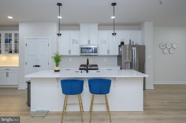 kitchen featuring white cabinets, glass insert cabinets, appliances with stainless steel finishes, hanging light fixtures, and a kitchen island with sink