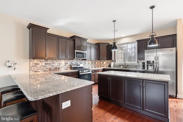 kitchen with dark wood finished floors, stainless steel appliances, hanging light fixtures, dark brown cabinets, and a kitchen bar