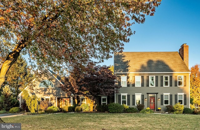 colonial-style house featuring roof with shingles, a chimney, and a front lawn