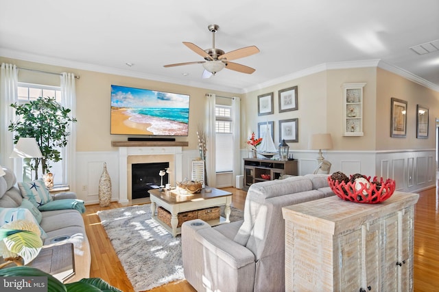 living area with visible vents, a wainscoted wall, a fireplace with flush hearth, ornamental molding, and wood finished floors