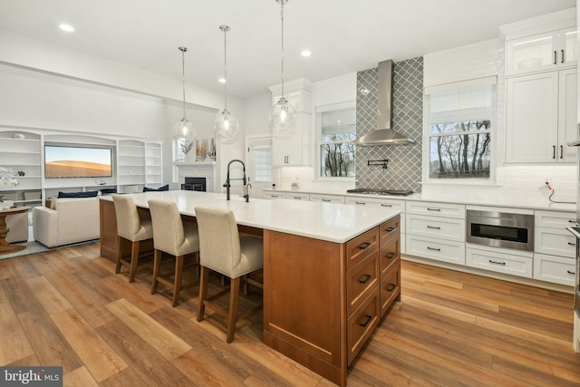 kitchen with white cabinets, an island with sink, wall chimney exhaust hood, open floor plan, and light countertops