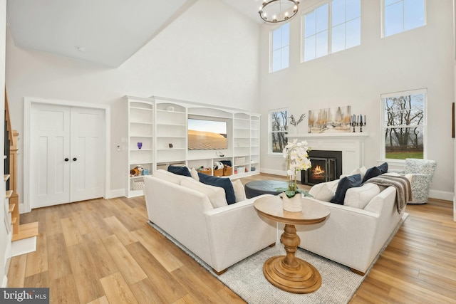 living room featuring a warm lit fireplace, a chandelier, light wood-style flooring, a towering ceiling, and baseboards