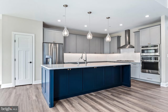 kitchen with stainless steel appliances, light countertops, wall chimney range hood, an island with sink, and pendant lighting