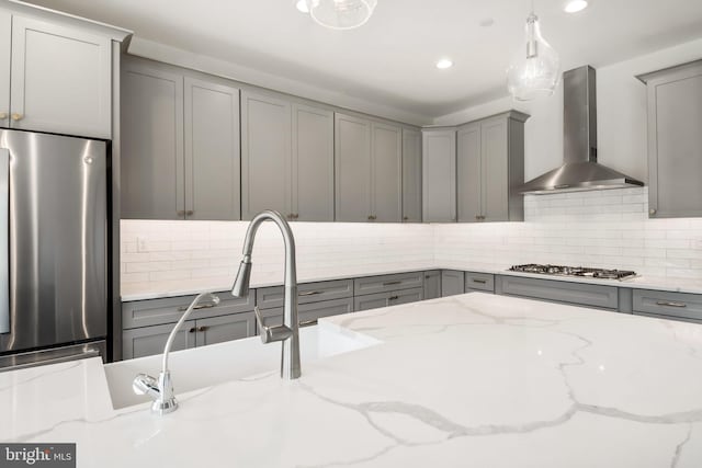 kitchen featuring light stone counters, pendant lighting, gray cabinets, appliances with stainless steel finishes, and wall chimney range hood