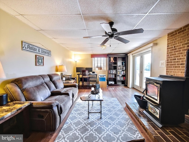 living area featuring a drop ceiling, a wood stove, a ceiling fan, and wood finished floors