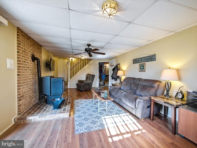 living room featuring ceiling fan, a drop ceiling, stairway, a wood stove, and wood finished floors