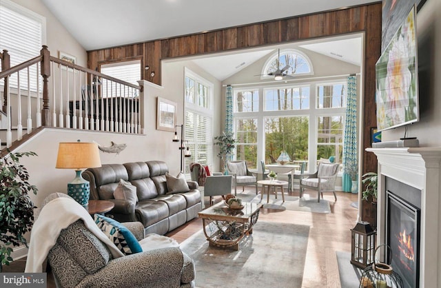 living room with wood finished floors, high vaulted ceiling, ceiling fan, stairs, and a glass covered fireplace