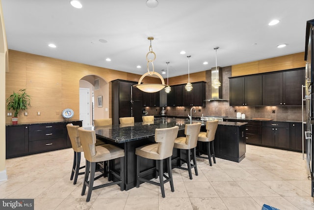 kitchen featuring arched walkways, a breakfast bar, a large island with sink, wall chimney range hood, and backsplash