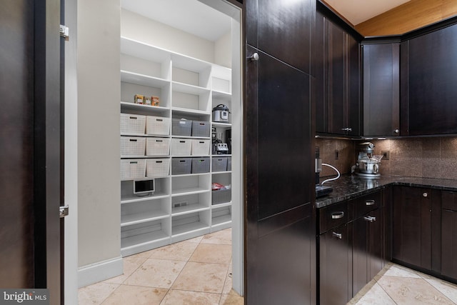 kitchen featuring light tile patterned floors, dark brown cabinets, dark stone countertops, and backsplash