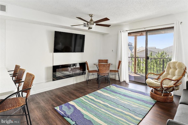 living room with a textured ceiling, ceiling fan, dark wood-style flooring, visible vents, and a glass covered fireplace