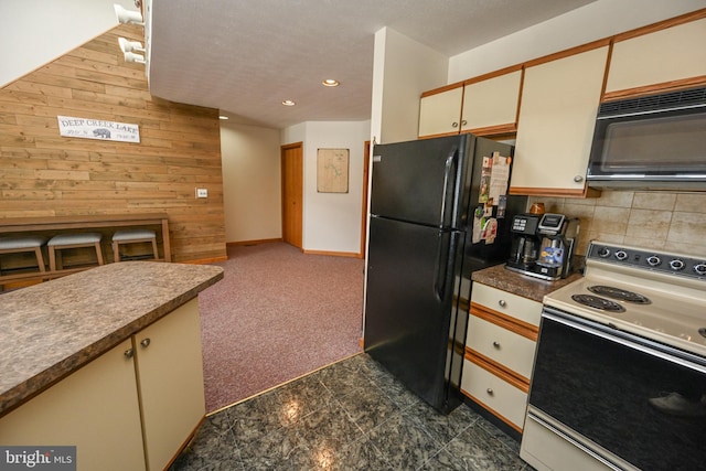 kitchen featuring wooden walls, decorative backsplash, cream cabinets, black appliances, and dark carpet