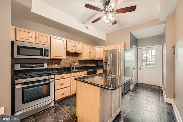 kitchen with light brown cabinetry, appliances with stainless steel finishes, a kitchen island, a sink, and dark stone countertops