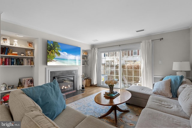 living room with built in shelves, a fireplace with flush hearth, wood finished floors, visible vents, and ornamental molding