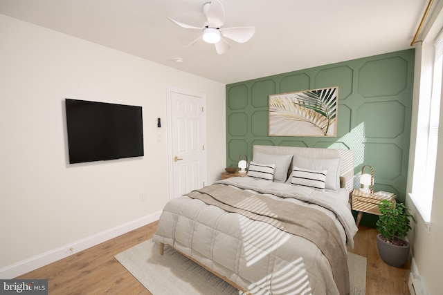 bedroom featuring ceiling fan, a baseboard heating unit, an accent wall, baseboards, and light wood-type flooring