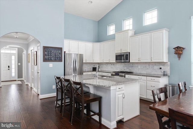 kitchen with arched walkways, light stone counters, stainless steel appliances, white cabinets, and a center island with sink