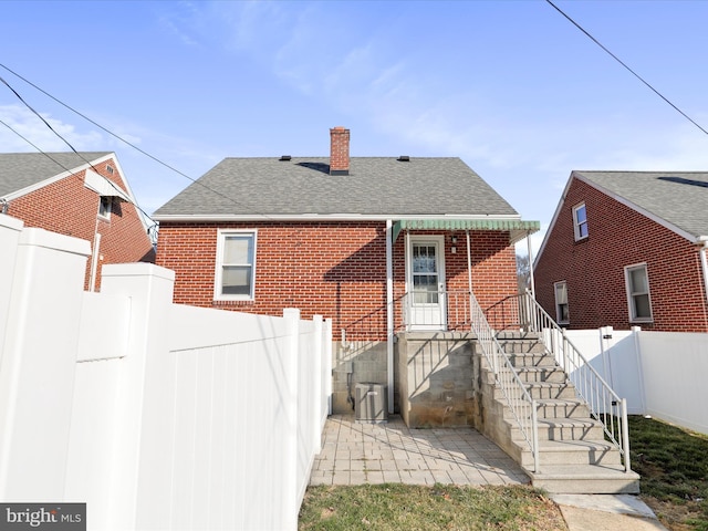 back of property featuring brick siding, a chimney, a shingled roof, and fence