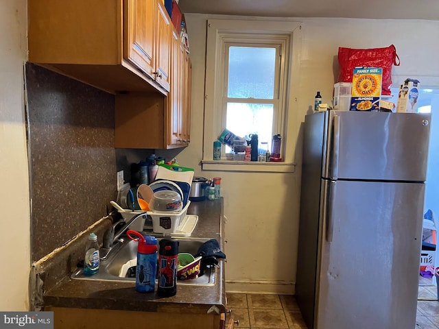 kitchen featuring dark countertops, brown cabinets, freestanding refrigerator, tile patterned flooring, and a sink