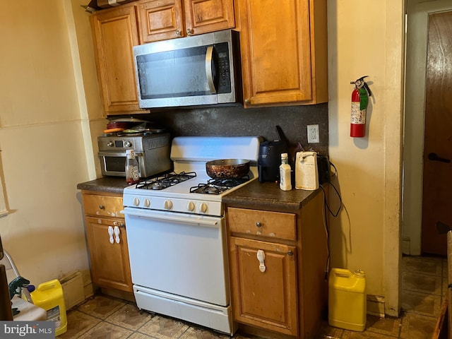 kitchen with brown cabinetry, dark countertops, stainless steel microwave, and white gas stove