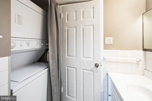 laundry room featuring laundry area, tile walls, a wainscoted wall, stacked washer / drying machine, and a sink