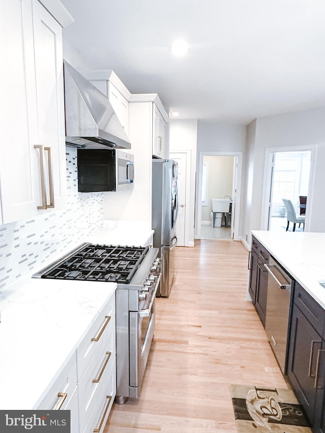 kitchen with tasteful backsplash, appliances with stainless steel finishes, white cabinetry, light wood-type flooring, and wall chimney exhaust hood