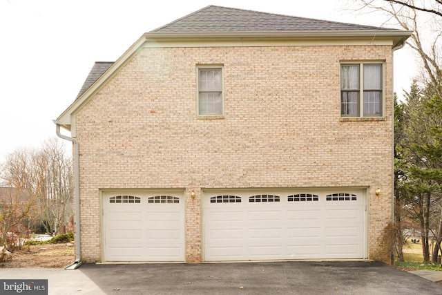 view of side of property featuring driveway, a shingled roof, an attached garage, and brick siding