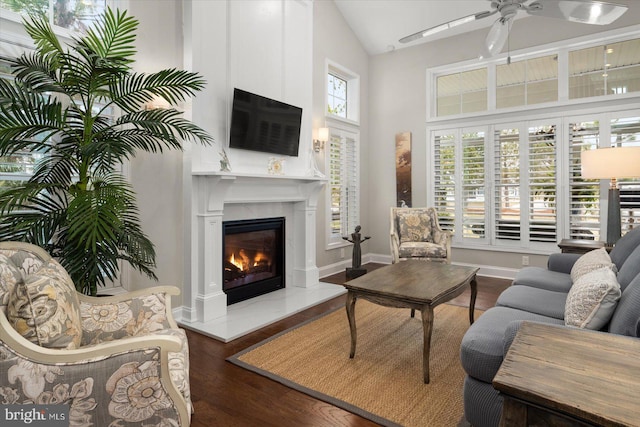 living area with high vaulted ceiling, a ceiling fan, wood finished floors, a glass covered fireplace, and baseboards