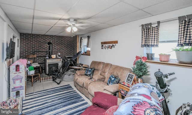 tiled living area featuring a wood stove, brick wall, a ceiling fan, and a paneled ceiling