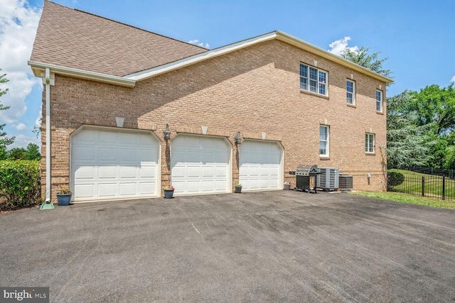 view of side of home with aphalt driveway, brick siding, roof with shingles, an attached garage, and central AC unit