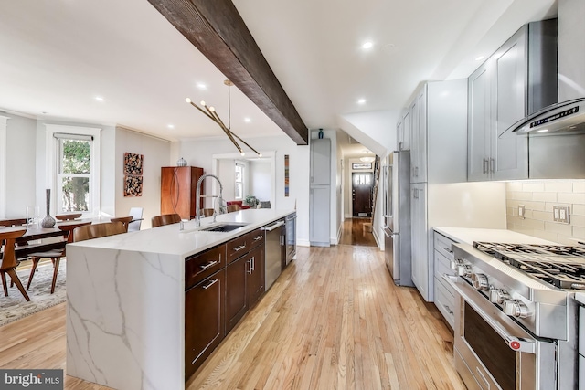 kitchen with appliances with stainless steel finishes, wall chimney range hood, an island with sink, light wood-type flooring, and beamed ceiling