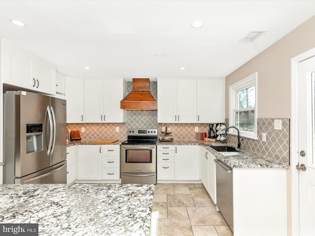 kitchen featuring appliances with stainless steel finishes, light stone counters, a sink, premium range hood, and backsplash