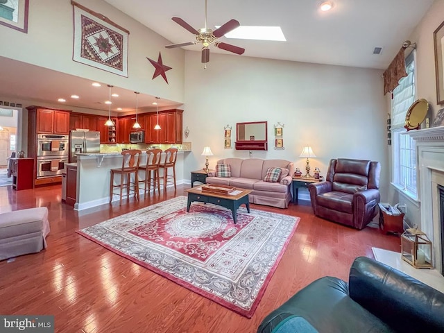 living area featuring a skylight, a fireplace with flush hearth, ceiling fan, wood finished floors, and high vaulted ceiling