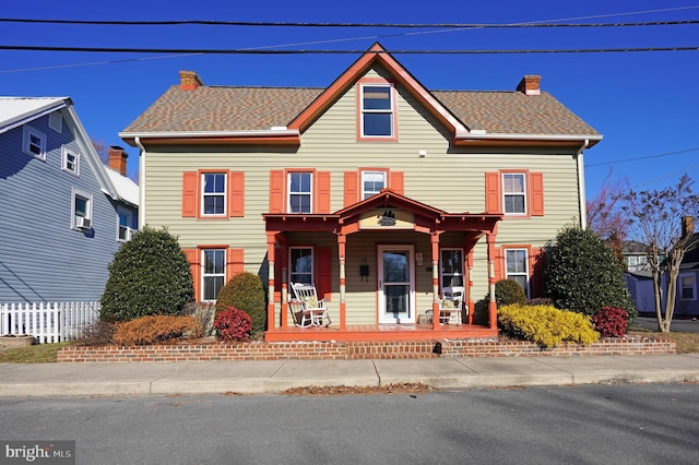 view of front facade with a porch, a shingled roof, a chimney, and fence