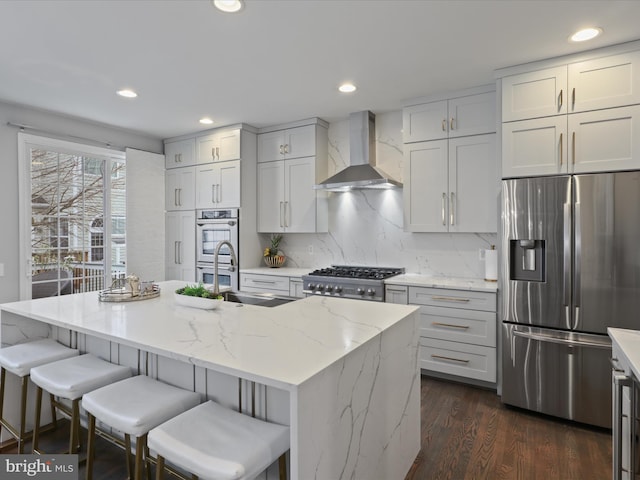 kitchen featuring appliances with stainless steel finishes, a kitchen island with sink, light stone counters, and wall chimney exhaust hood