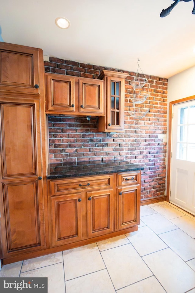 kitchen with light tile patterned flooring, brick wall, brown cabinets, dark stone counters, and glass insert cabinets