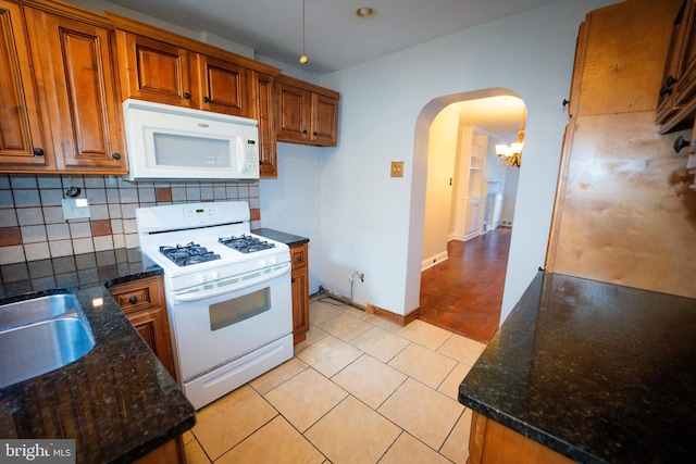 kitchen featuring arched walkways, brown cabinets, tasteful backsplash, dark stone countertops, and white appliances