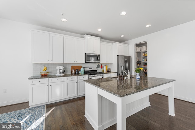 kitchen with dark wood finished floors, appliances with stainless steel finishes, white cabinets, a kitchen island with sink, and a sink