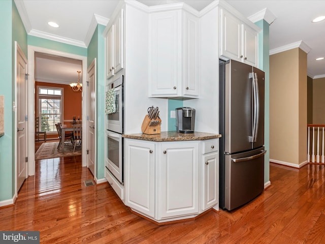 kitchen with light wood-style flooring, appliances with stainless steel finishes, ornamental molding, white cabinetry, and dark stone counters