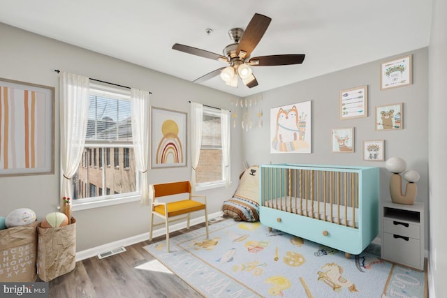 bedroom with a crib, baseboards, visible vents, a ceiling fan, and wood finished floors