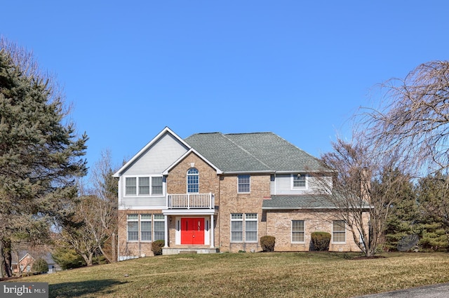 view of front of property featuring a balcony, brick siding, a front lawn, and roof with shingles