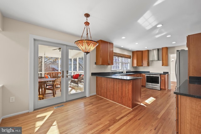 kitchen featuring a peninsula, appliances with stainless steel finishes, wall chimney exhaust hood, light wood finished floors, and dark countertops