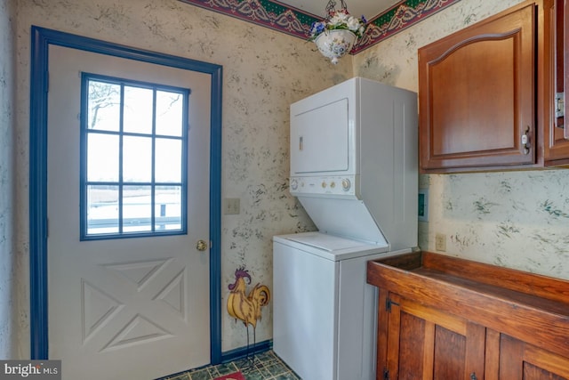 clothes washing area featuring stacked washer and dryer, a wealth of natural light, cabinet space, and wallpapered walls
