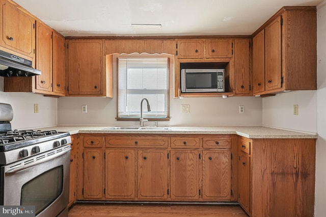 kitchen with light countertops, appliances with stainless steel finishes, brown cabinetry, a sink, and under cabinet range hood